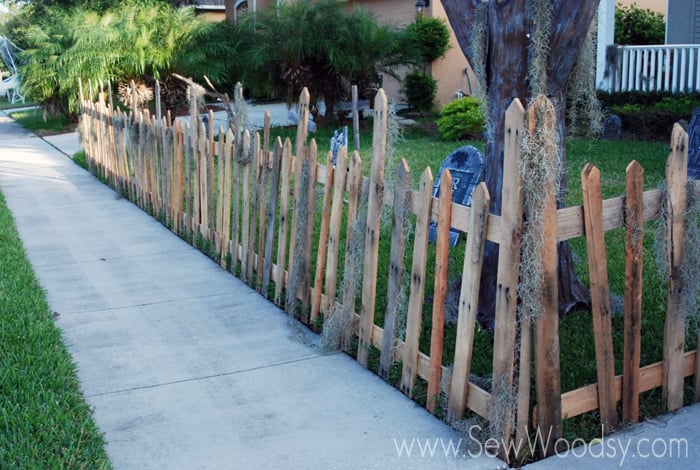 Wood fence lining a side walk with moss hanging on the fence and gravestones in the background. 