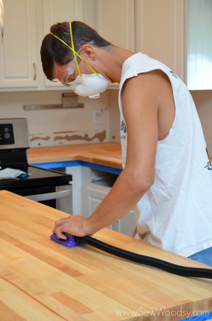 Man in white cutoff shirt wearing dust mask and safety goggles sanding Butcher Block Countertops.