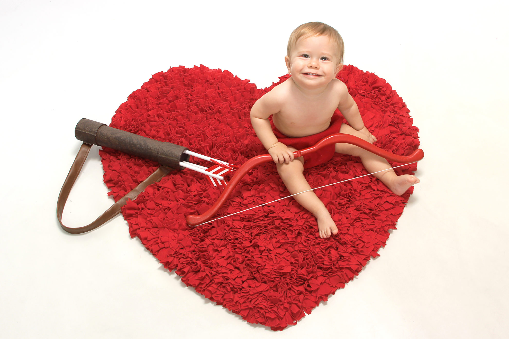 Red heart rug with a baby dressed as cupid sitting on top.