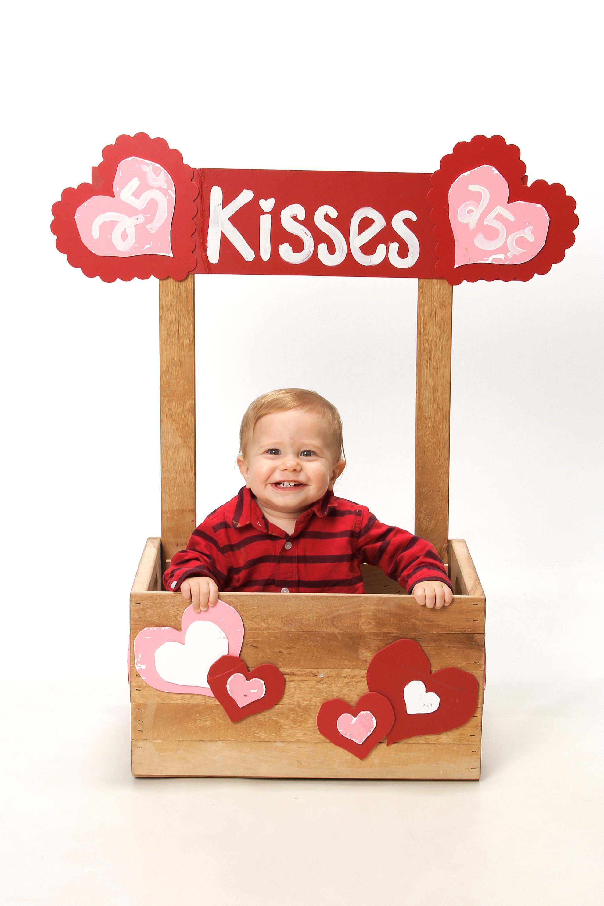 Little baby boy sitting in a kissing booth made out of a wood crate with hearts stapled on front.