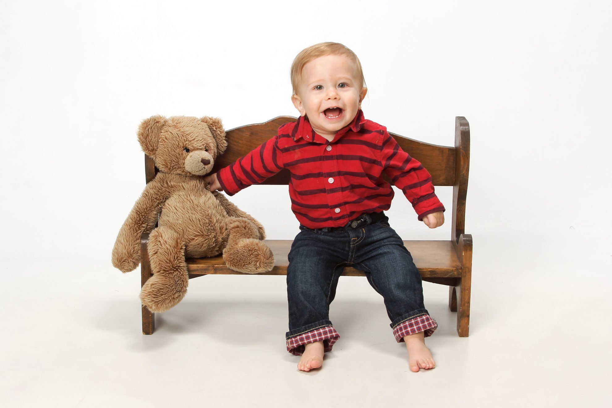 Little baby boy sitting on a wooden bench with a teddy bear next to him.