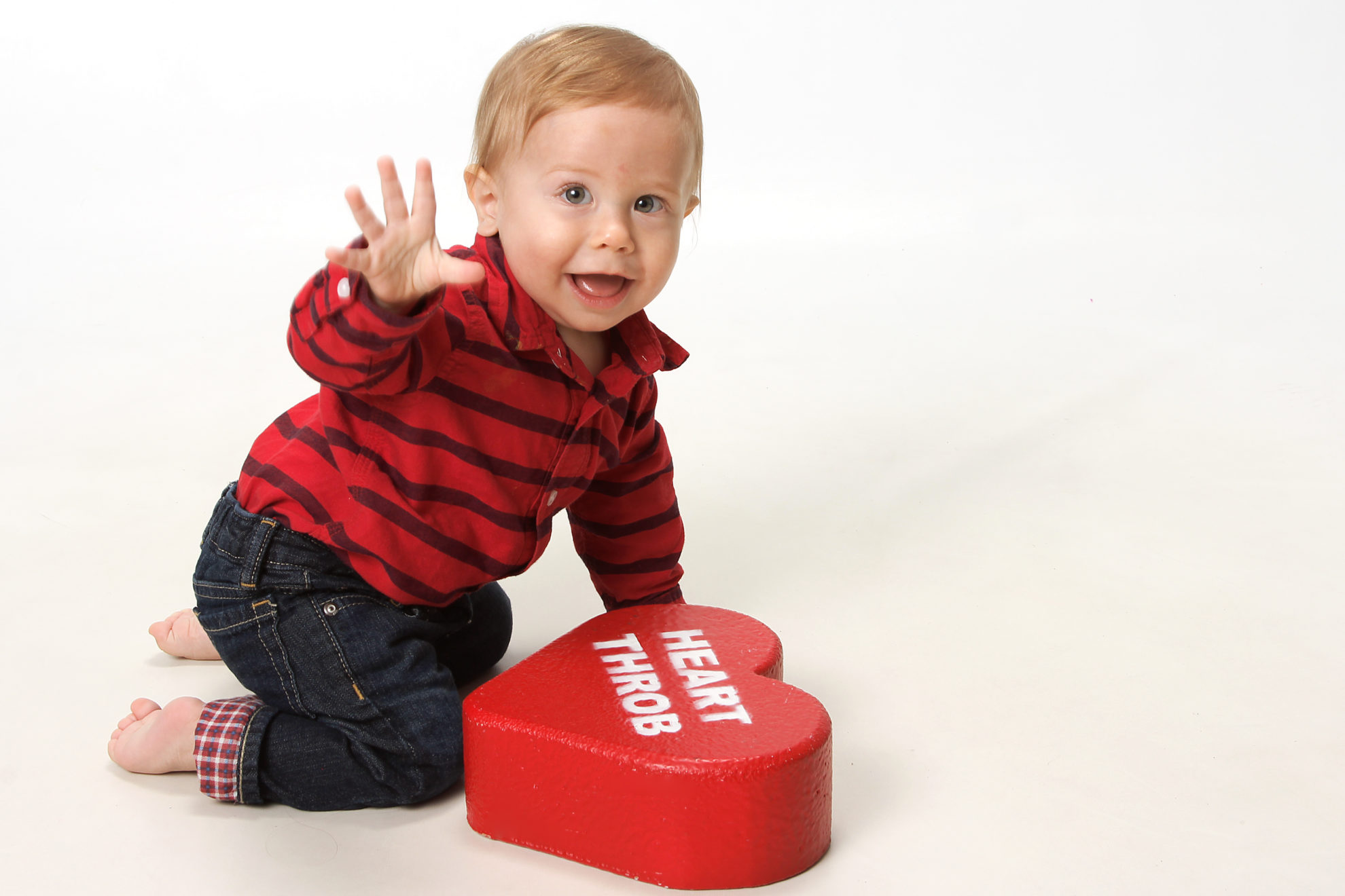 Little boy baby on knees waving at the camera with other hand on a heart throb heart.