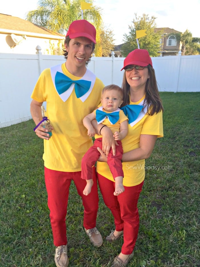 Male, female, and baby dressed as Tweedledum and Tweedledee  standing in a green yard with white fence.