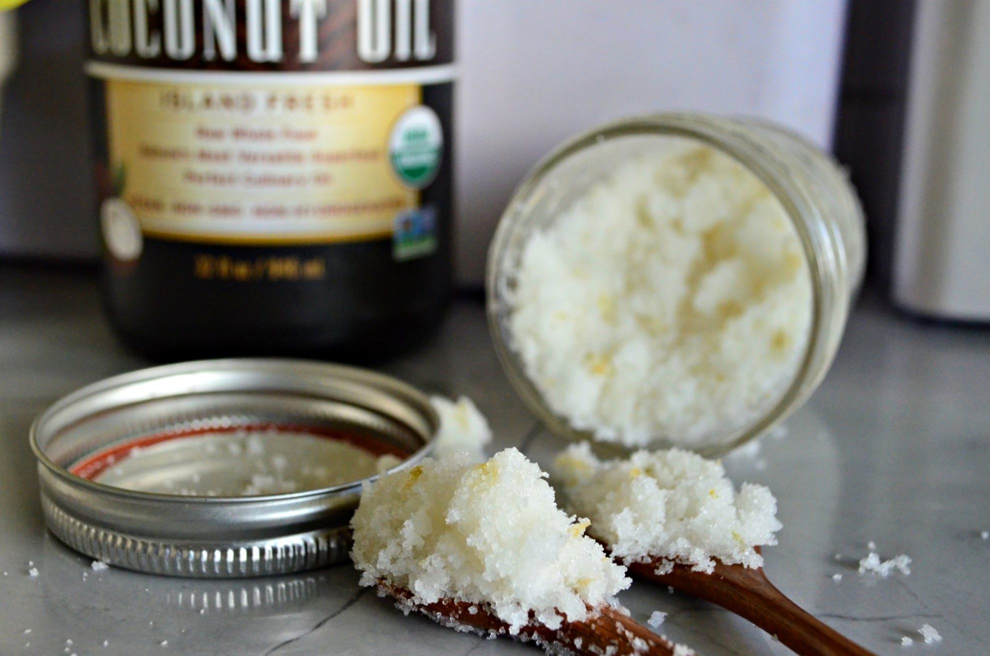 Glass jar on the side with sugar scrub coming out of it with sugar scrub on two wooden spoons on a countertop.