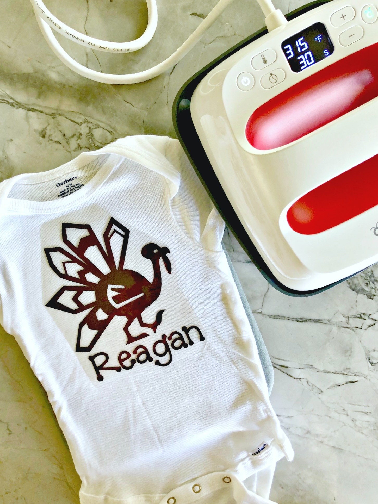 Top view of a white onesie with a turkey and name iron on vinyl cut out next to an EasyPress.