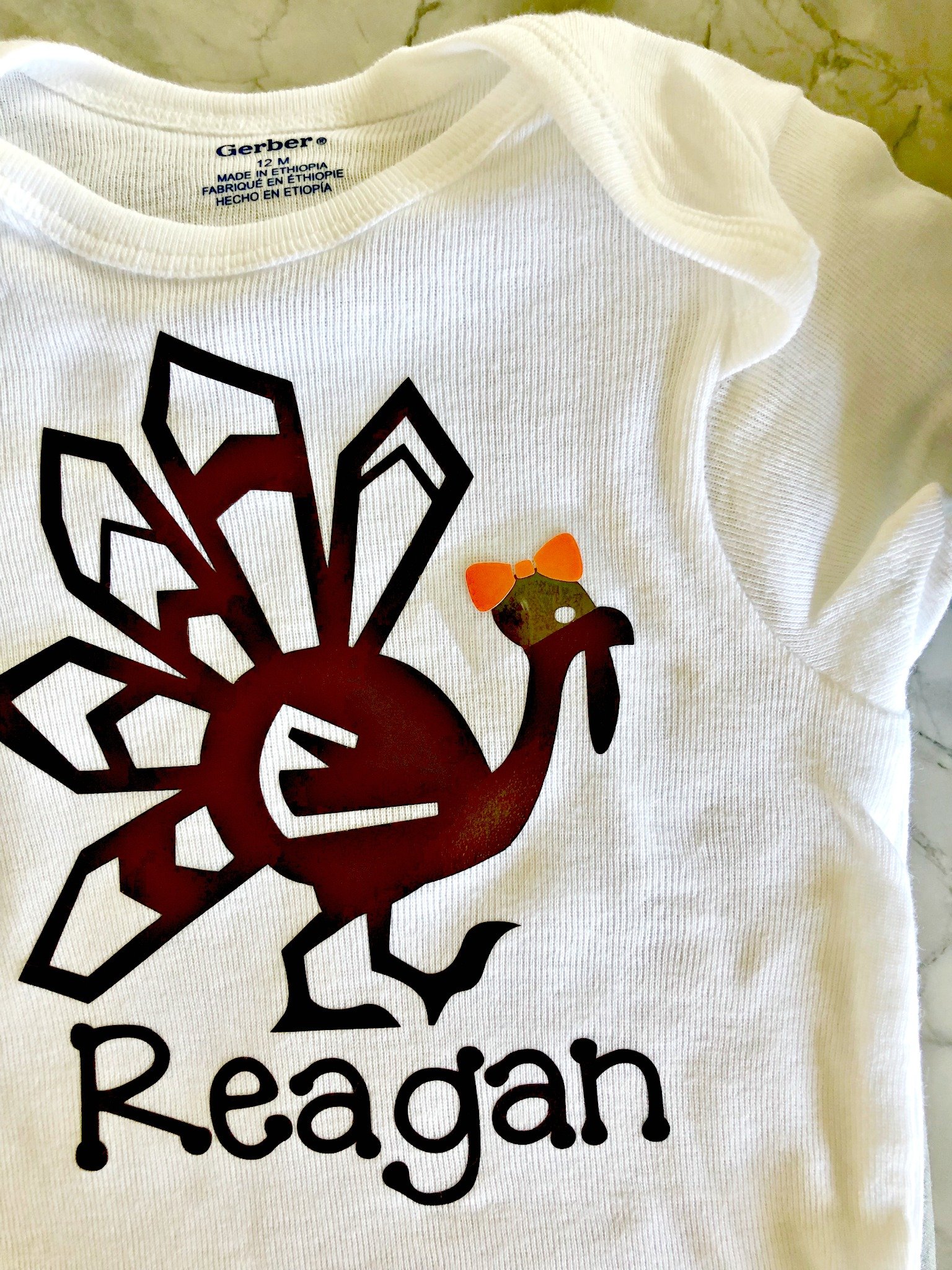 White onesie with a brown turkey, name, and bow being applied on it. 