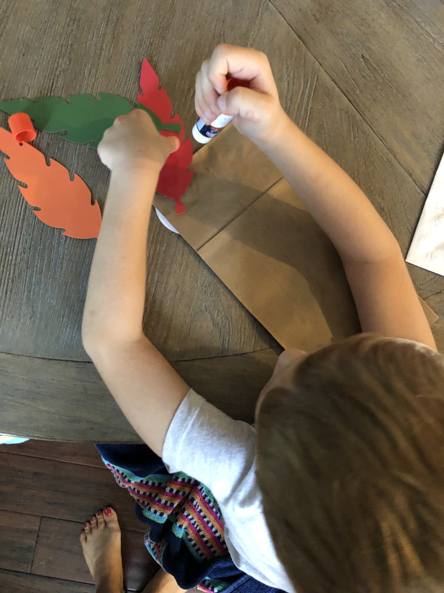 Top view of a little boy glueing feathers on a back of a brown paper bag.