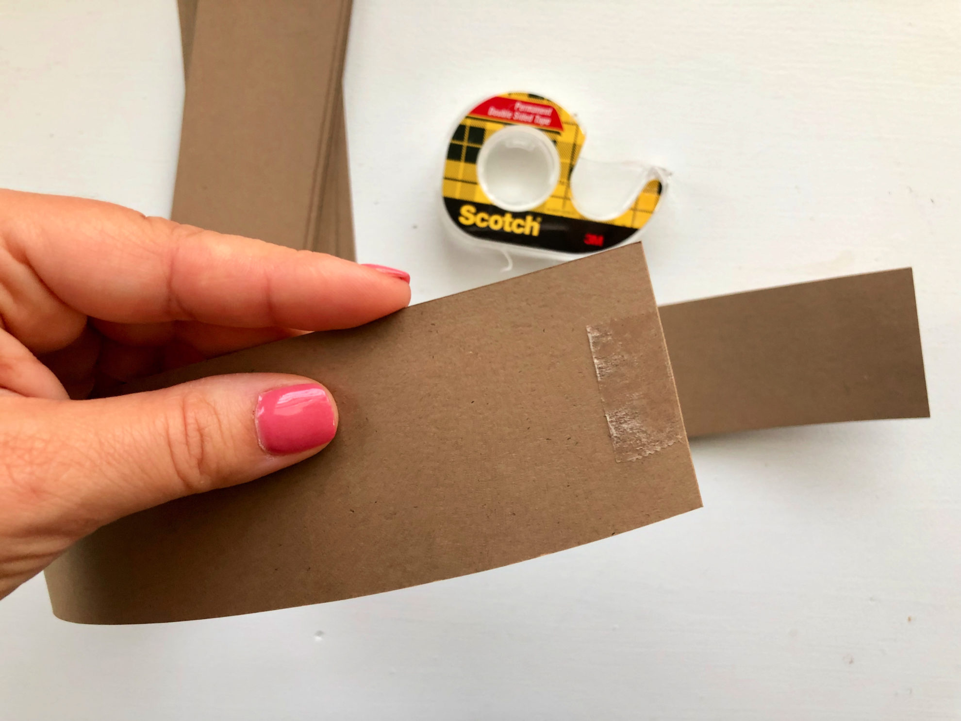 Female hand with pink fingernails holding brown cardstock strip with double sided tape on it.