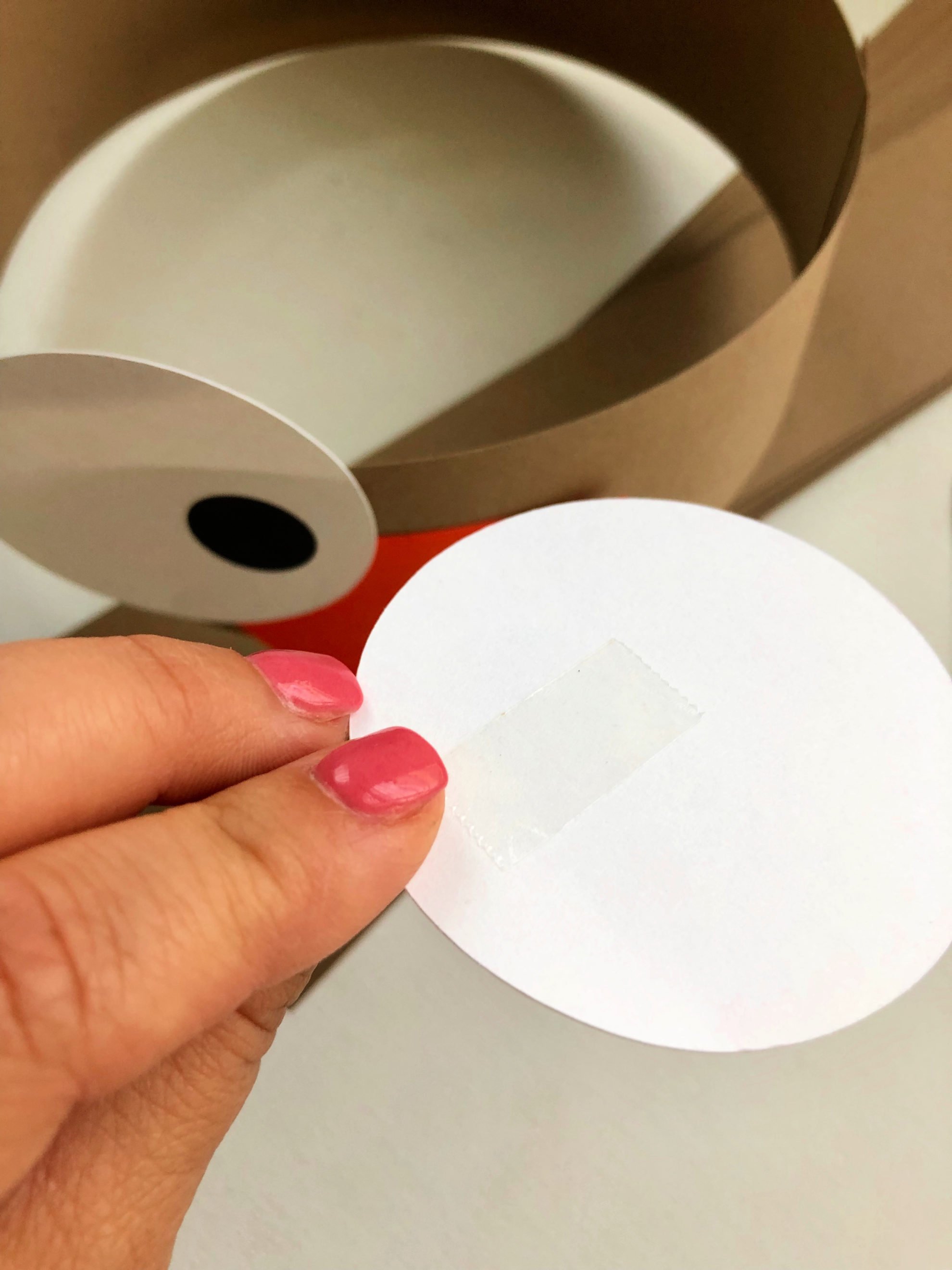 Female hand with pink nails holding a white circle with double stick tape on it.