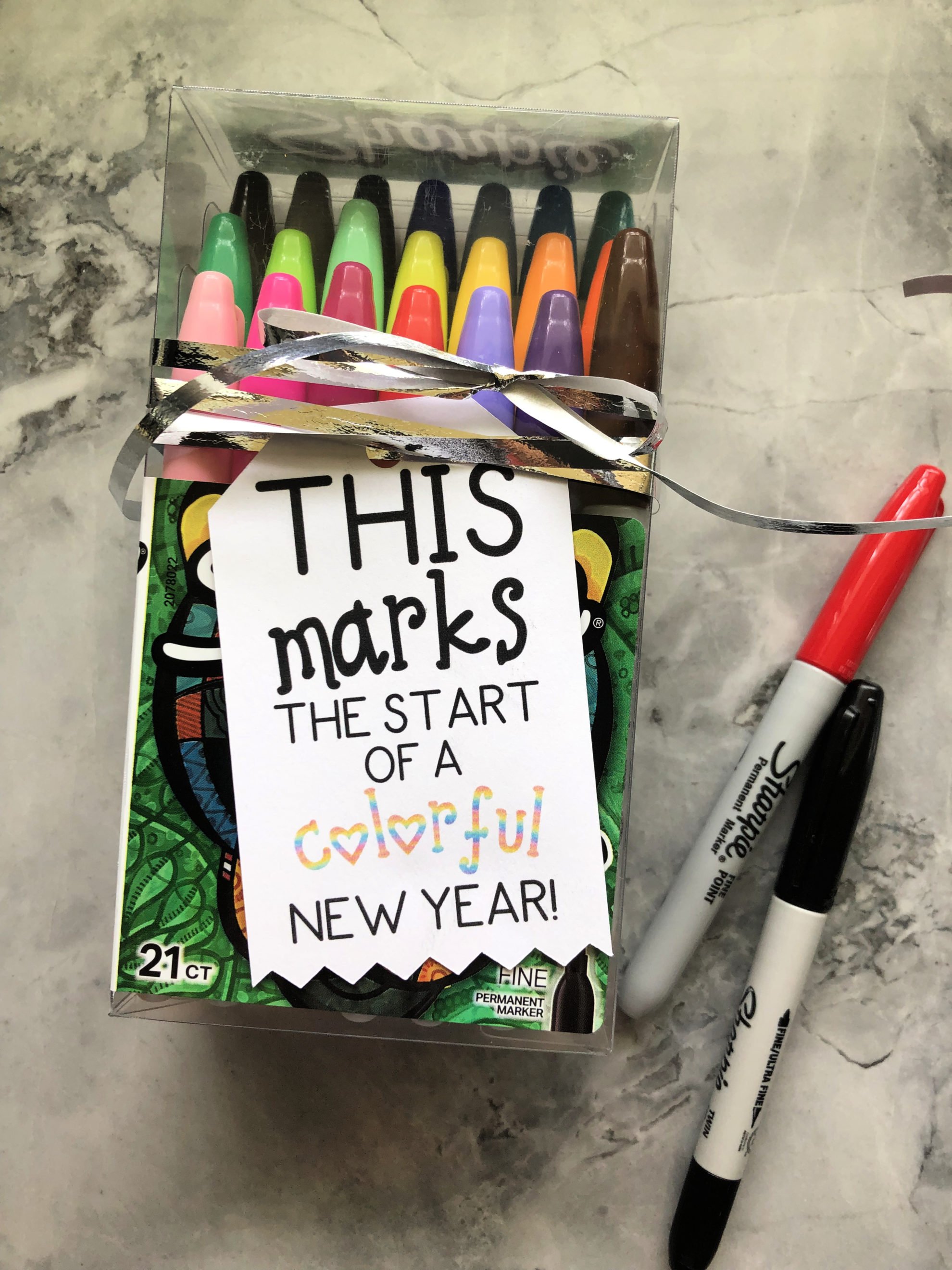 Box of sharpies with a tag and silver ribbon on it resting on a marble countertop. 