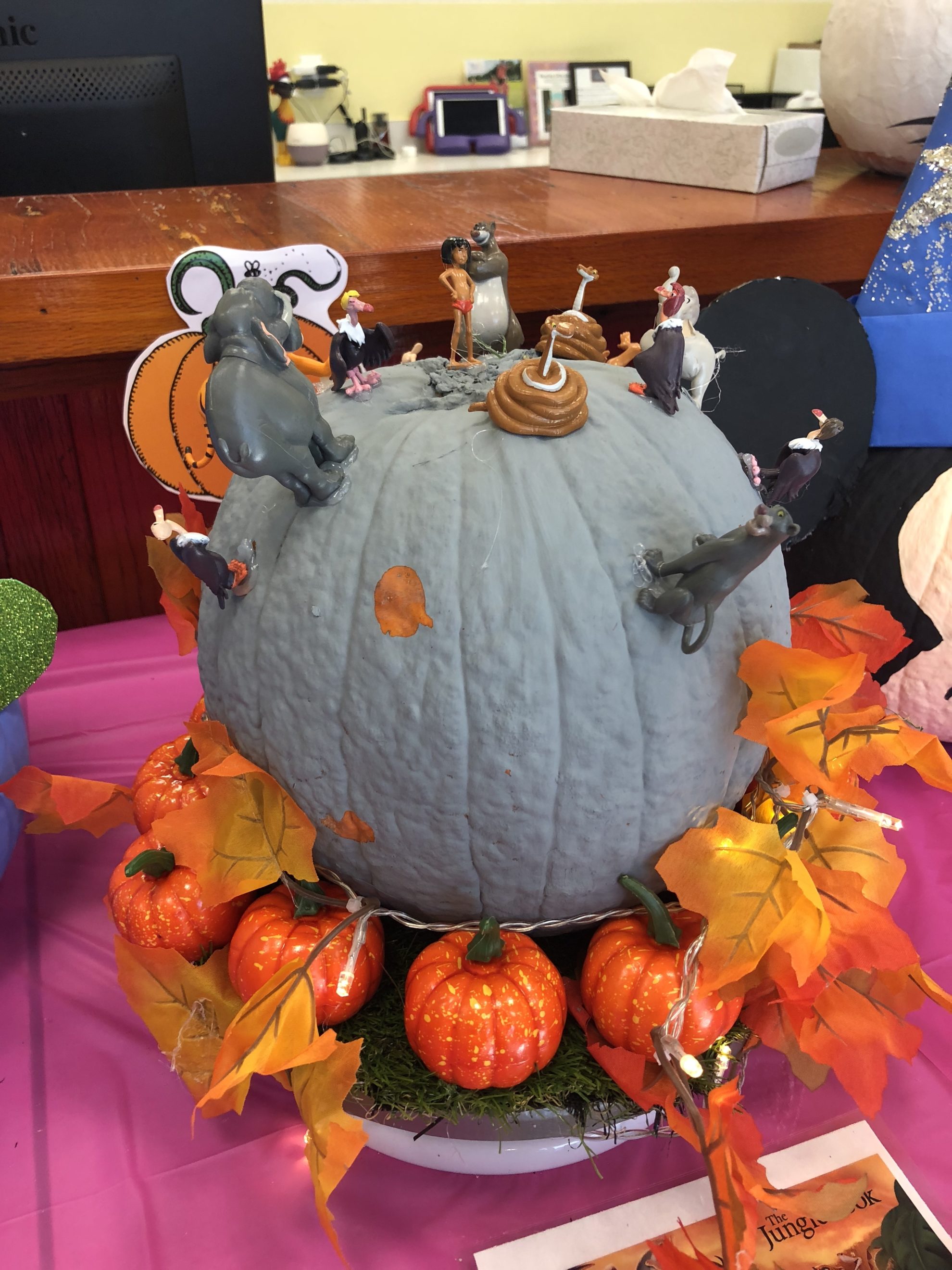 Grey painted pumpkin with The Jungle Book characters on top with leaves and pumpkins around it. 