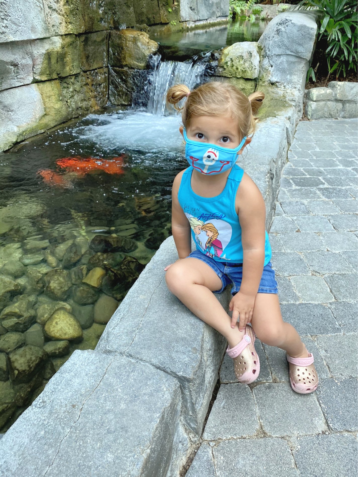 Little girl with blue mask on face sitting next to a koi pond. 