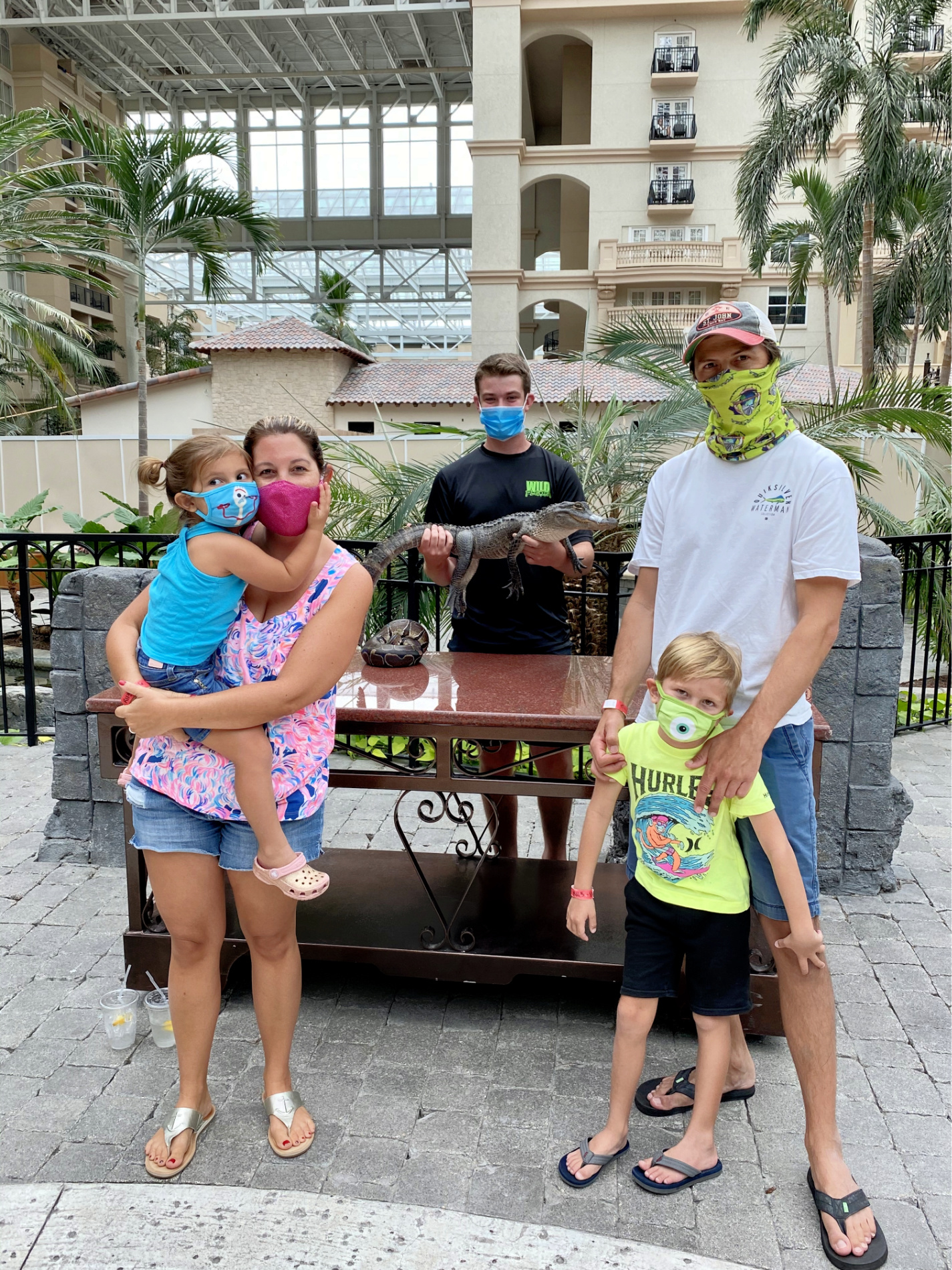 Family standing with masks with man with mask holding alligator and ball python on table.