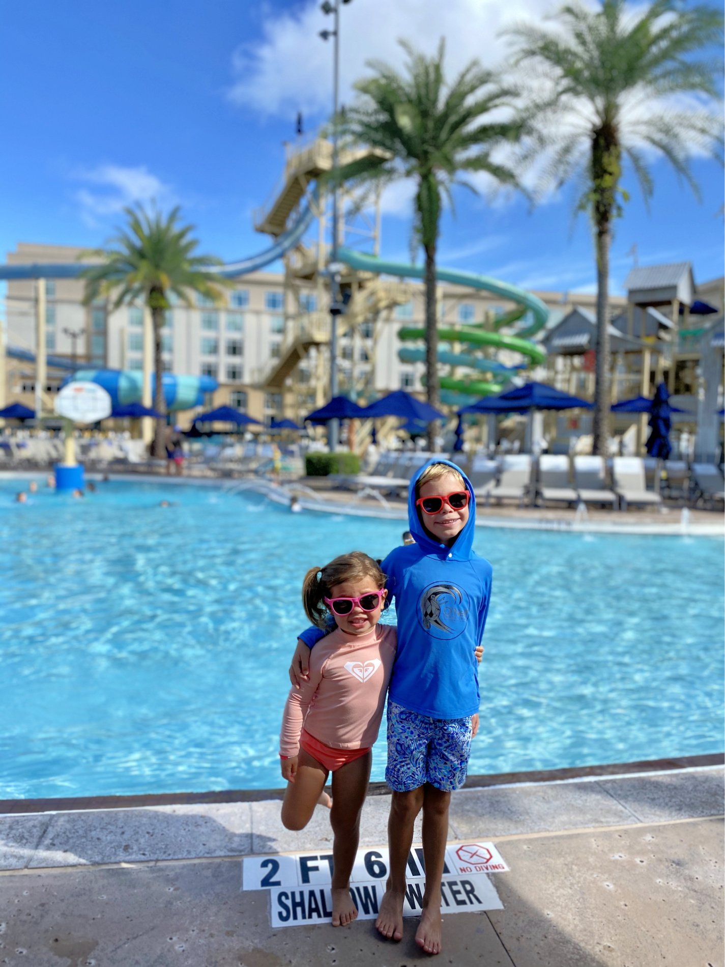 Two children in swimsuits and sunglasses standing in front of a swimming pool.