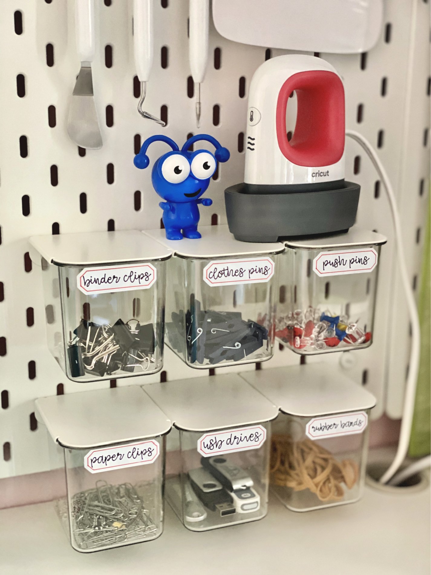 Clear bins on a peg board with hand written labels on them with a blue cricket and EasyPress Mini.