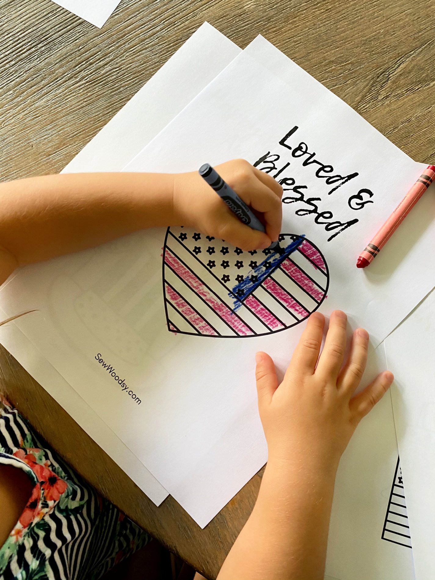 Top view of a little girl coloring an American Flag heart with text on paper that reads "Loved & Blessed".