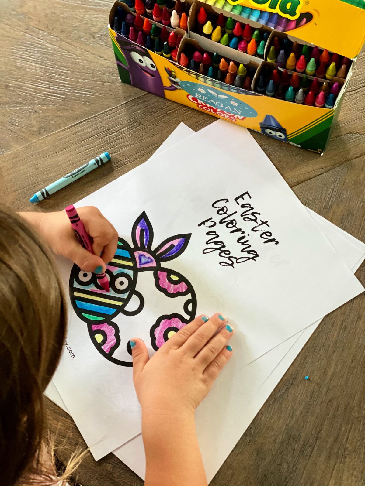 Little girl with blue fingernails coloring an Easter coloring page with crayons.