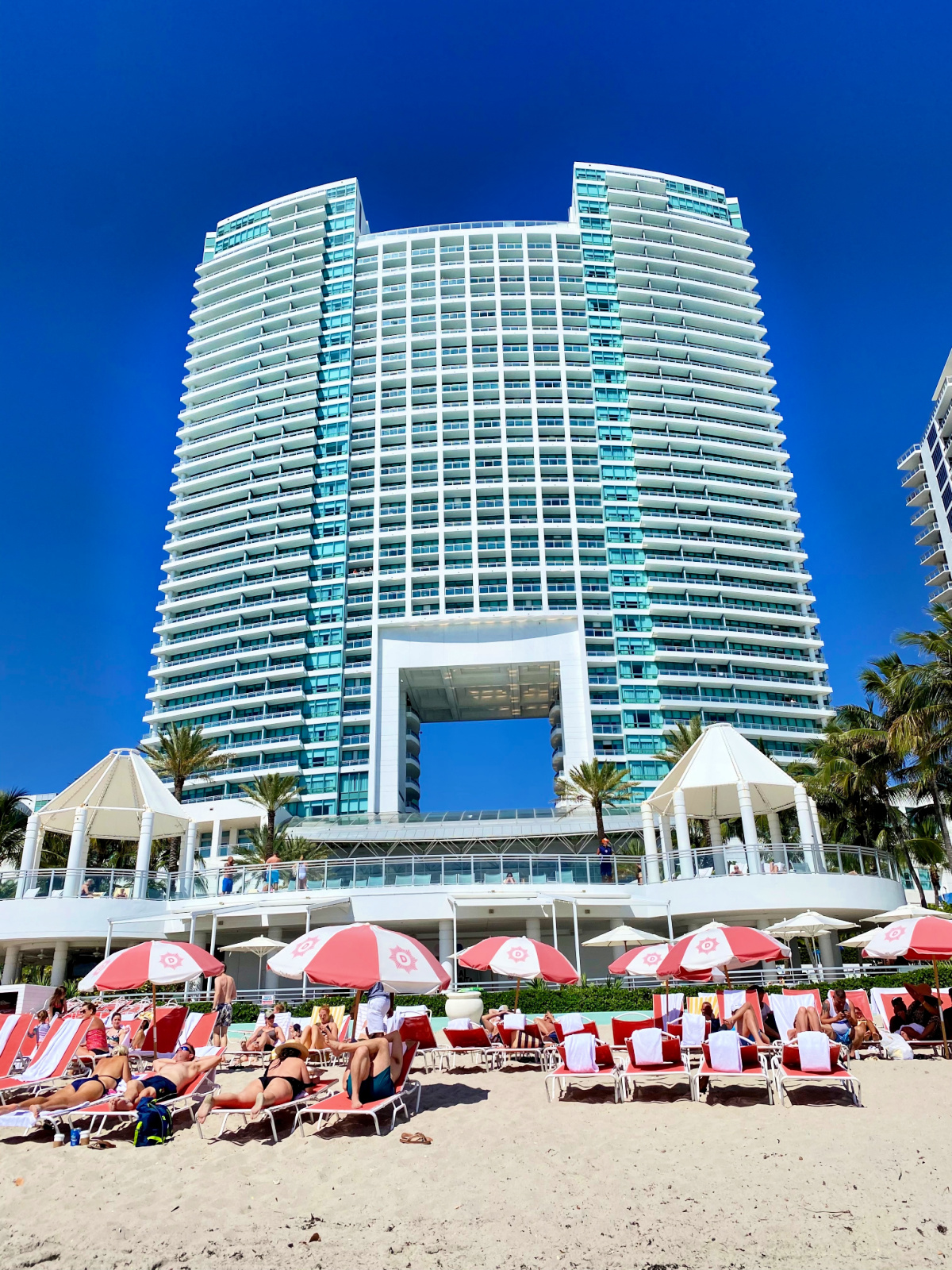 Large hotel on the beach with sunbathers on chairs and umbrellas.