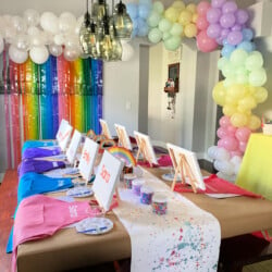 Pastel rainbow balloon arch with long brown table with aprons, easels, canvases, beads, and placemats.