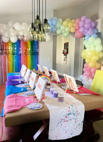 Pastel rainbow balloon arch with long brown table with aprons, easels, canvases, beads, and placemats.