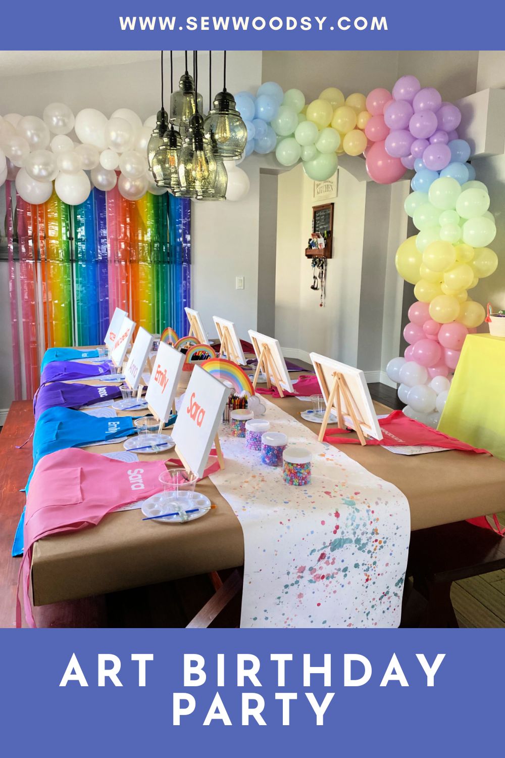 Table with aprons, canvas and a rainbow balloon arch in the background.