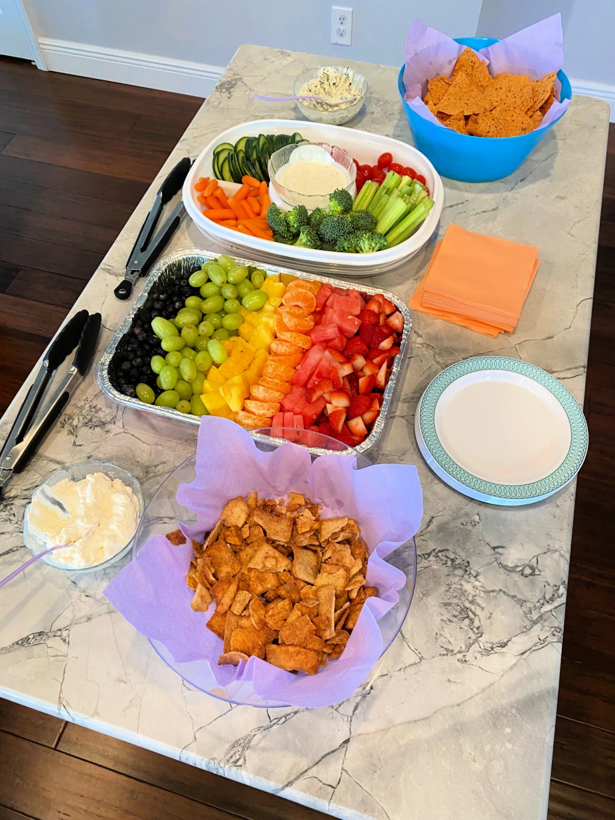 Rainbow fruit tray, veggie tray, chips and dip on marble kitchen island.