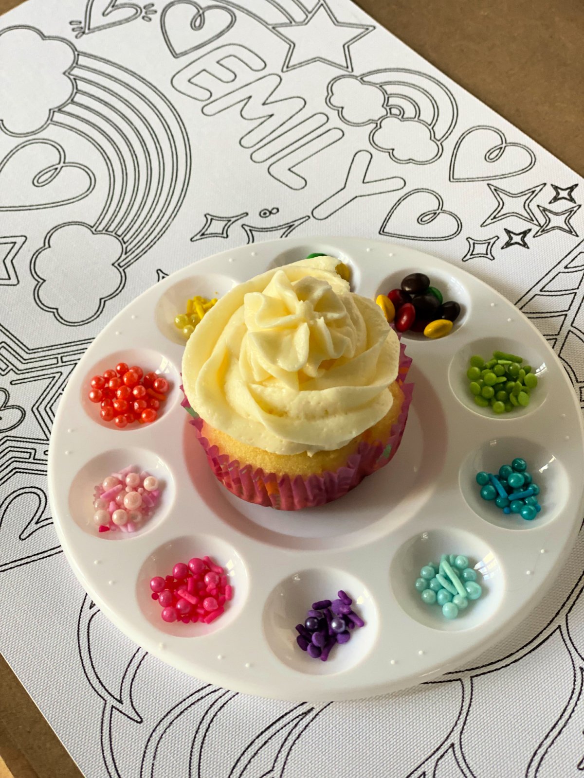 Close up of a paint palette filled with sprinkles with a vanilla frosted cupcake.