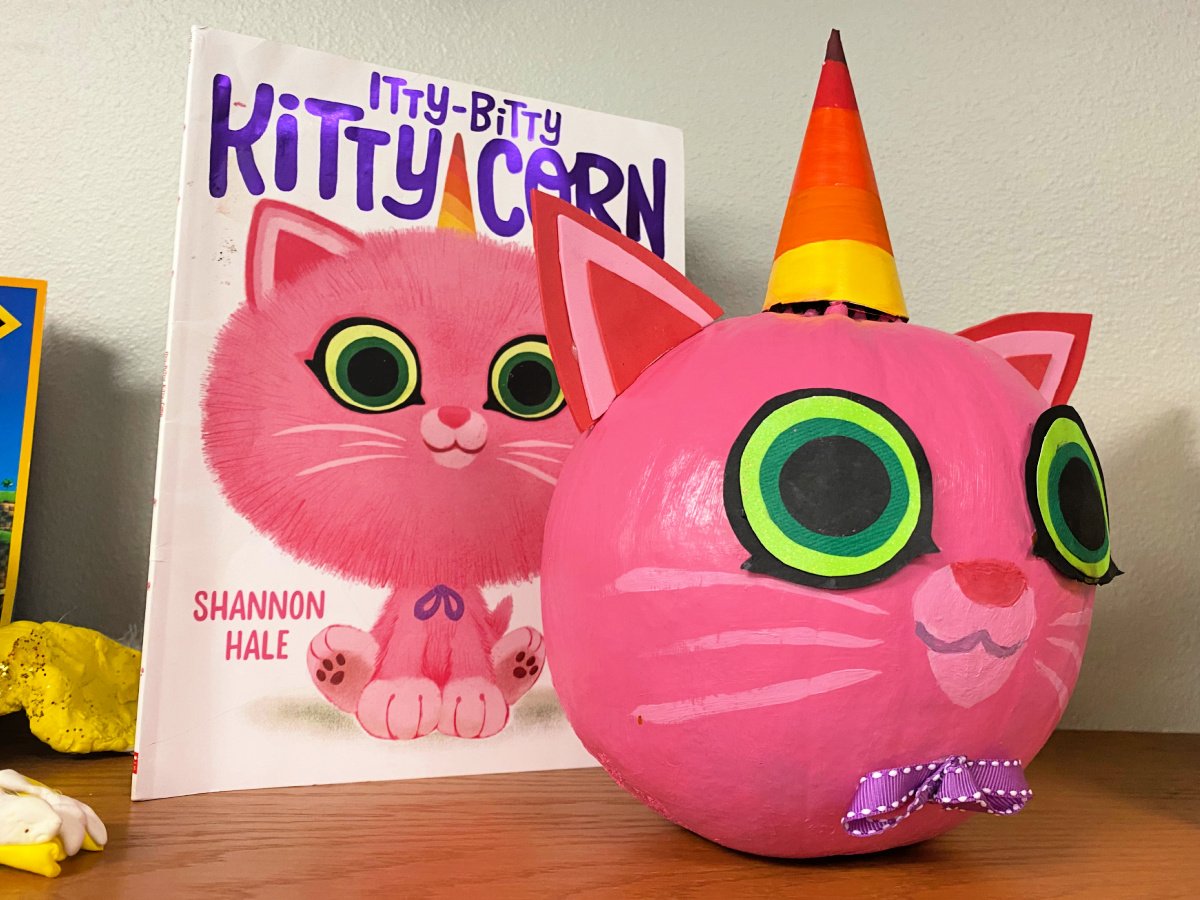 Pink pumpkin painted as a kitty with a candy corn horn on top next to a book called Itty-bitty kitty corn.