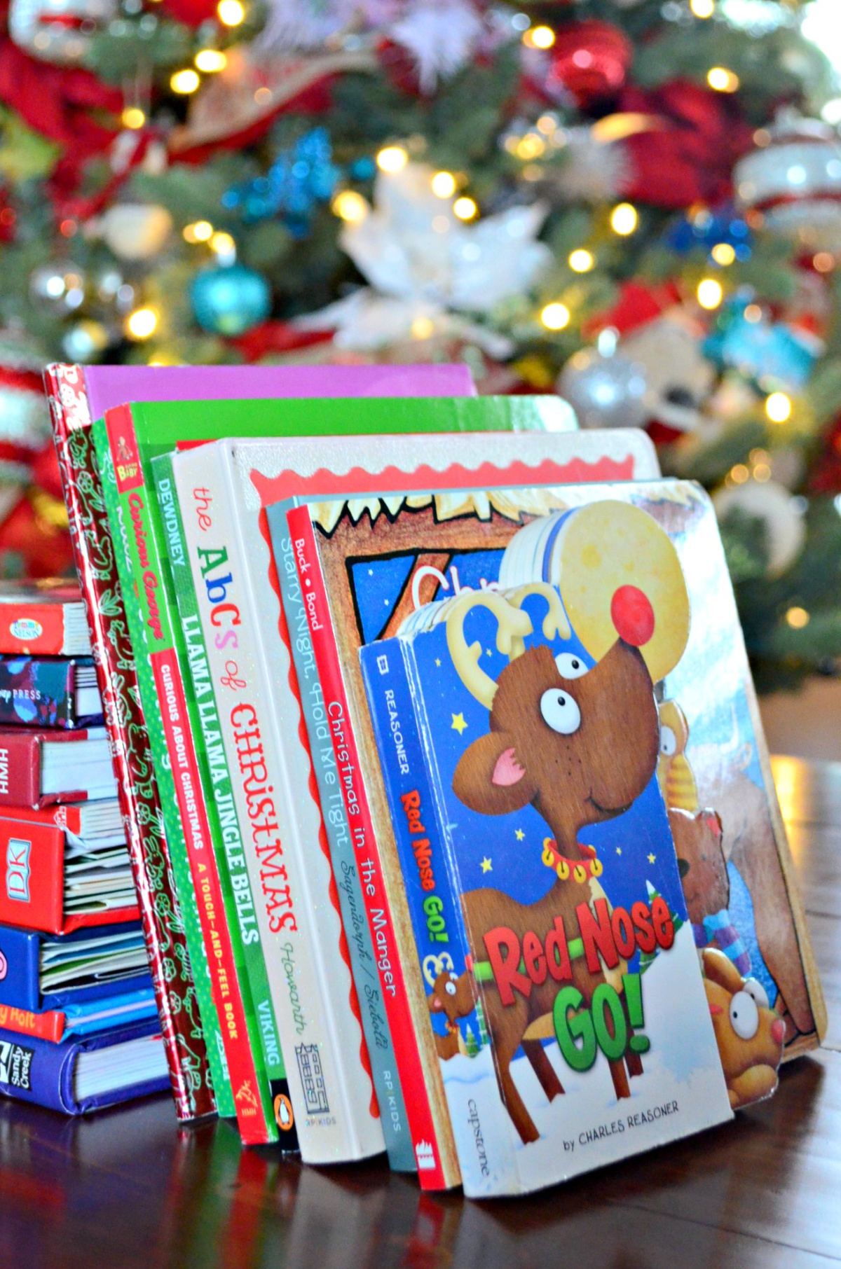 A stack of Christmas books on a wood table with a christmas tree in the background.