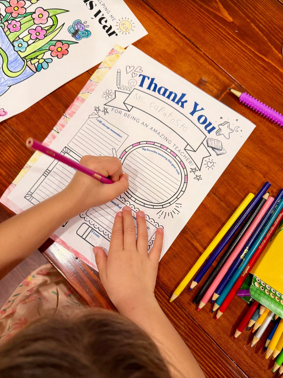 Little girl filling out a thank you teacher printable using a colored pencil on a wood table.